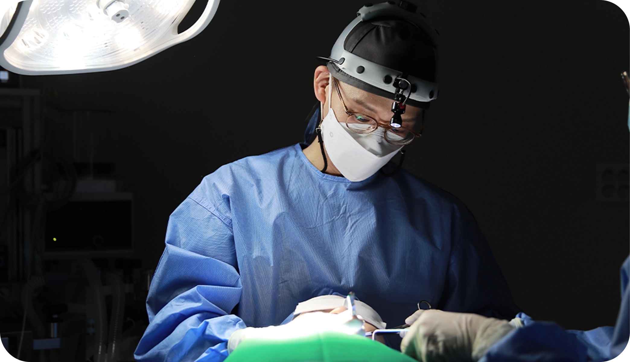 手術中の医師。ライト付きヘッドギア、マスク着用。345 Plastic Surgeryは美容整形、特に顔の整形手術に特化。高度な技術で患者の理想を追求。関連キーワード：美容整形、顔の整形手術。